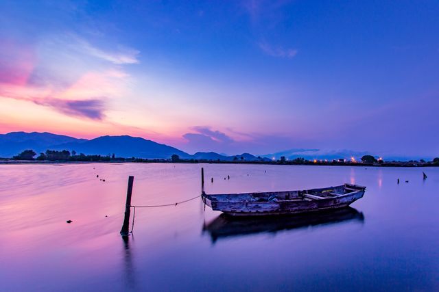 Old fishing boat moored on a tranquil lake with still water and a mountain backdrop at sunset. Cozy evening ambiance characterized by colorful twilight sky and peaceful reflections on the water's surface. Ideal for travel blogs, relaxation visuals, meditation themes, and nature lovers.