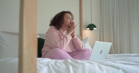 African american woman sitting contemplative on bed using laptop in pink loungewear