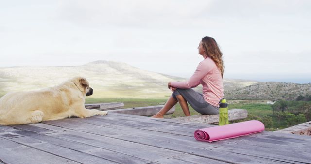 Woman practicing yoga outdoors on a wooden platform with dog nearby. Scenic landscape in background with gentle hills and clear sky. Great for themes of wellness, relaxation, meditation, and connection with nature. Suitable for websites, blogs, advertisements, and wellness content aiming to evoke tranquility and harmony.