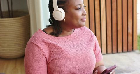 African american woman listening with headphones holding smartphone on patio near doorway