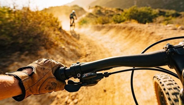 Gloved hand steering mountain bike handlebar on dusty sunlit off-road trail at golden hour