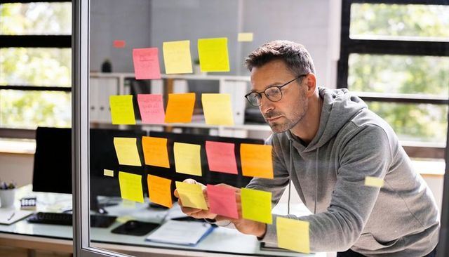 Focused man leaning over glass partition adjusting colorful sticky notes for agile planning