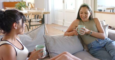 Two diverse women relaxing on sofa drinking from mugs and chatting in modern living room