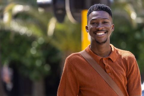 Smiling african man standing on sunlit city street wearing rust sweater and crossbody bag