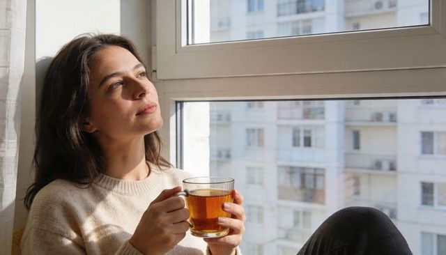 Woman sipping tea while gazing out sunlit apartment window, cozy urban morning