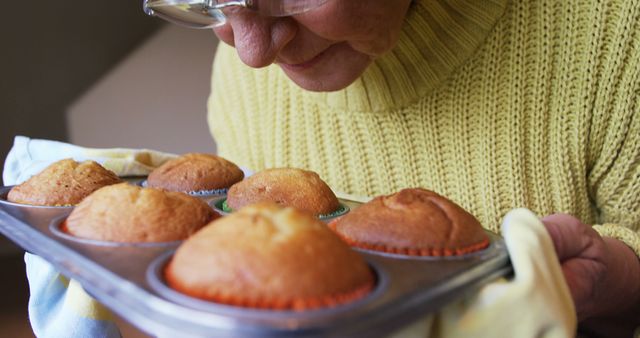 A senior woman is examining freshly baked muffins in a tray, with copy space. Her attention to detail suggests a passion for baking and the warmth of homemade cuisine.