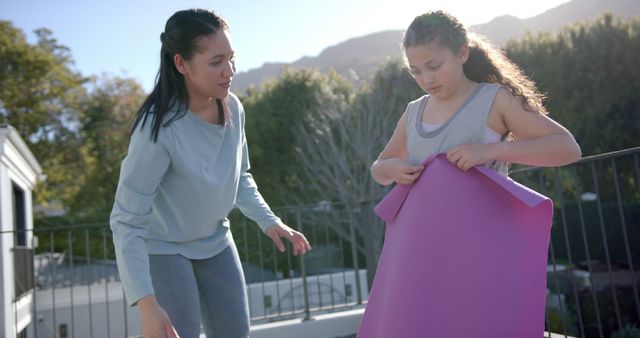 Mother is helping her daughter prepare a yoga mat for an outdoor exercise session on a sunny day. They are on a terrace or balcony, trees and mountains in background. This image can be used for articles on family fitness, yoga, bonding activities, and promoting healthy lifestyles for children.