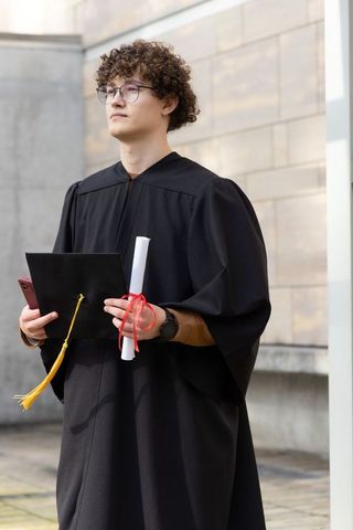 Young man in twenties holding diploma and mortarboard on campus celebrating graduation