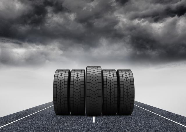 Five tires are neatly aligned on an asphalt road with a dramatic, stormy sky in the background. This image can be used for automotive advertisements, tire promotions, safety campaigns, and articles related to road trips or vehicle maintenance. The dark clouds and overcast weather add a sense of urgency and drama, making it suitable for themes related to weather challenges and road safety.