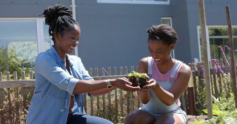 African woman and teen girl planting seedlings, bonding in sunny backyard garden
