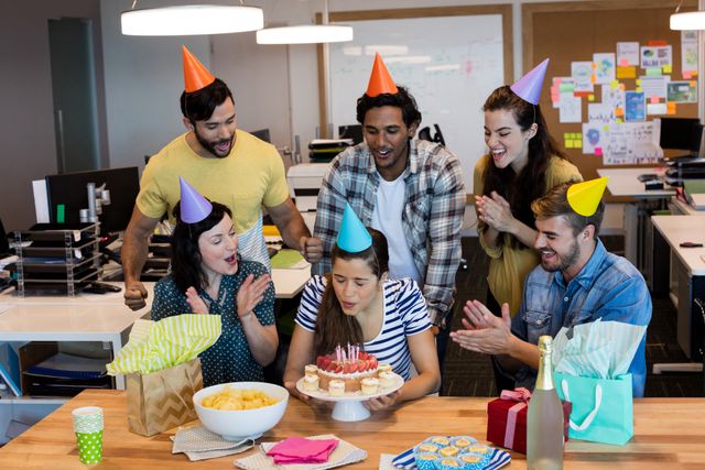 Group of colleagues celebrating a team member's birthday in an office environment. The birthday person is blowing out candles on a cake while others cheer and clap. Party hats, gifts, and snacks are present, creating a festive atmosphere. Ideal for use in articles or advertisements about workplace culture, team building, employee appreciation, and corporate events.