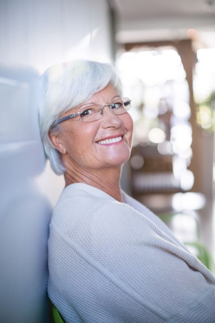 Senior woman with white hair and glasses sitting on sofa in clinic, smiling warmly. Ideal for healthcare, wellness, and senior care themes. Can be used in brochures, websites, and advertisements promoting medical services, senior living, and patient care.