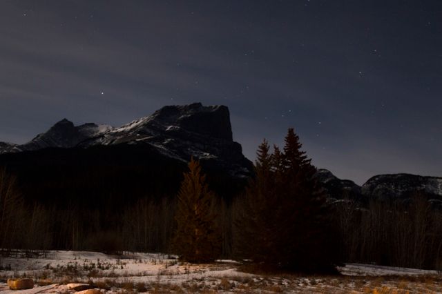 Majestic mountain range silhouetted against star-filled night sky with trees in foreground. This serene and tranquil winter scene is perfect for use in nature and travel blogs, adventure publications, outdoor-themed websites, or as a calming background for presentations.