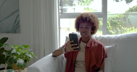 African american woman relaxing on sofa using smartphone near sunlit window with plant