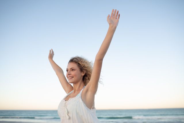 Woman standing on beach with arms raised, smiling joyfully. Ideal for concepts of freedom, happiness, relaxation, and vacation. Perfect for travel promotions, lifestyle blogs, wellness articles, and advertisements.