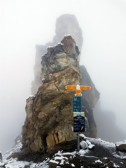 Trail signpost set against a towering snow-covered rocky formation in misty mountain environment. Ideal for illustrating outdoor adventures, hiking guides, travel brochures, and alpine expedition concepts.