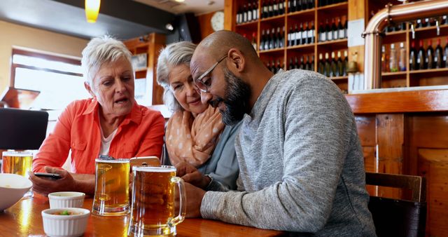 Group of diverse friends, including two senior women and a middle-aged man, socializing and enjoying beers at a cozy pub. They are engaged in conversation and interacting with their smartphones. Suitable for themes related to friendship, social interactions, leisure activities, seniors, and relaxation.