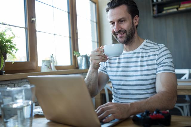 Smiling man using laptop and having a cup of coffee in coffee shop