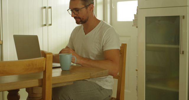 Man sitting at wooden table working on laptop with coffee mug beside, showcasing modern work-from-home lifestyle. Ideal for articles on remote work, productivity tips, technology use in daily life, or home office setups.