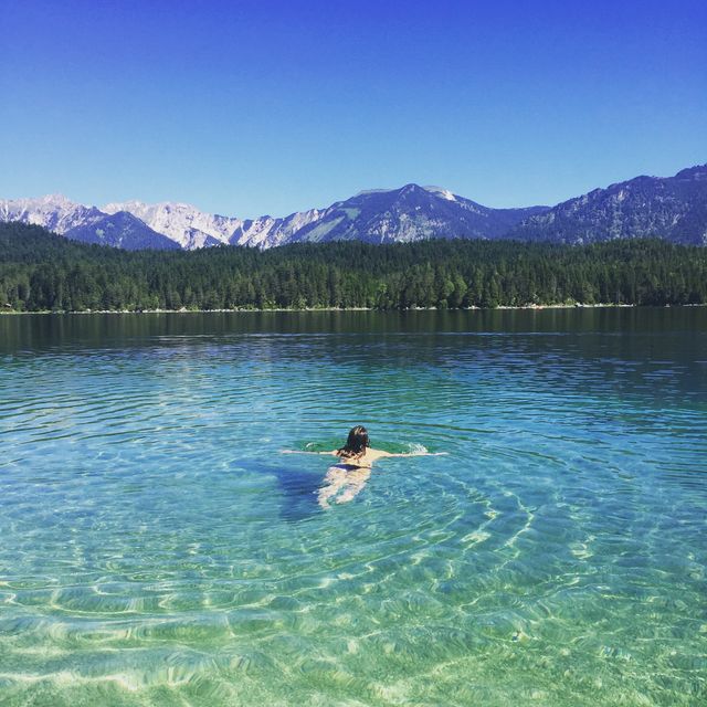 Woman enjoying swim in clear mountain lake surrounded by scenic forest and mountains. Ideal for promoting travel, nature adventures, summer activities, and wellness vacations.