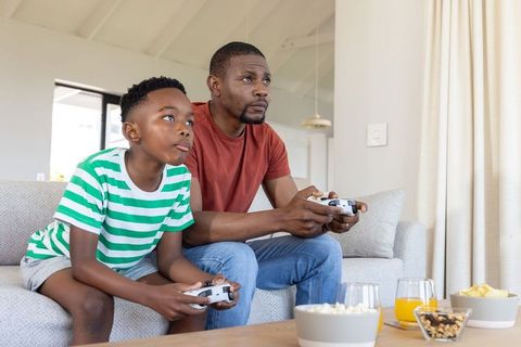 African american father and son playing console games on sofa, sharing popcorn and juice