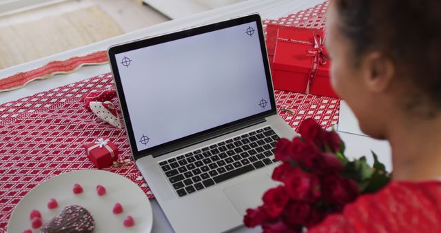 Woman holding bouquet of red roses while video calling on laptop, surrounded by Valentine's Day gifts, chocolates, and decorations. Ideal for Valentine's Day promotions, romantic greeting cards, or online advertisements for virtual celebrations.