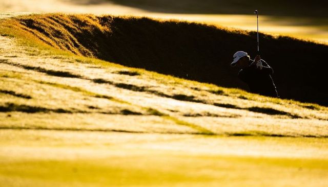 Golfer swinging from bunker rim during golden hour - dramatic silhouette and textured fairway