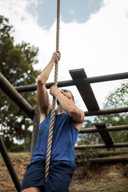 Fit man climbing rope in outdoor obstacle course, showcasing strength and determination. Ideal for fitness, outdoor training, and adventure-themed content. Perfect for promoting boot camps, physical challenges, and workout routines.