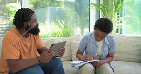African american father and teen daughter sharing learning on couch with tablet and notebook