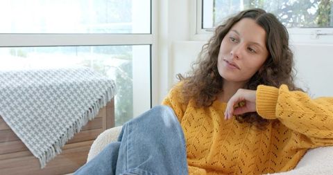 Woman in mustard sweater gazing through window, relaxing in sunlit cozy living room