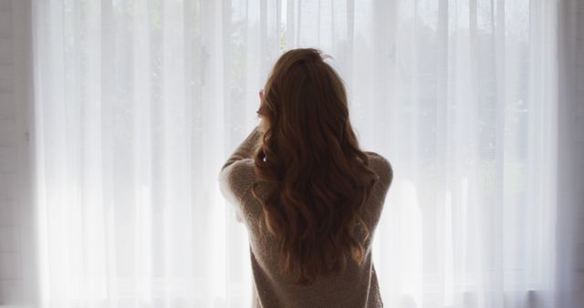 Caucasian woman standing at window with white curtain at home. Domestic life and lifestyle, unaltered.