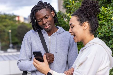 Two female friends sharing smartphone and laughing on urban rooftop in casual hoodies