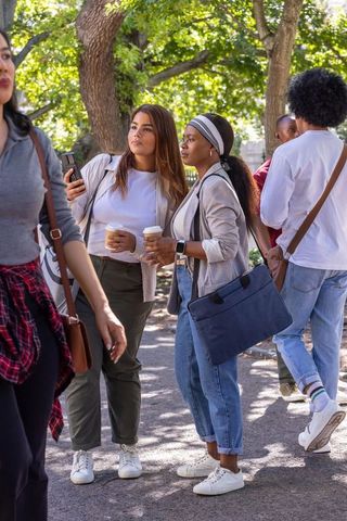 Hispanic and african american women holding coffee, checking phone on campus path