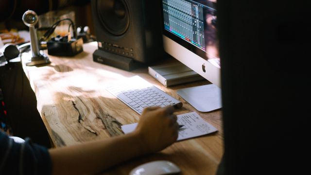 Music producer working at home studio, using computer and audio equipment for sound mixing and editing. Suitable for use in blog posts, articles, or advertisements focused on music production, remote workspaces, and creative professions.