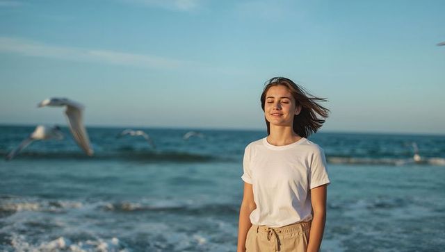 Standing woman facing ocean at sunlit beach, eyes closed, breeze blowing, seagulls flying