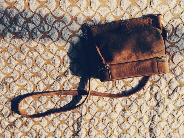 Brown leather handbag with shoulder strap placed on patterned rug under sunlight. Ideal for use in fashion blogs, online boutiques, product showcases, or commercial advertising targeting women's fashion and accessories. Can highlight details of quality leather product in both casual and formal contexts.