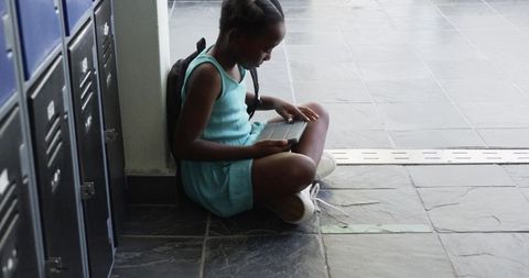 African girl sitting in school hallway studying on tablet beside lockers, tapping screen