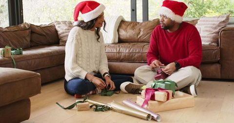 Diverse couple wrapping holiday gifts on living room floor wearing santa hats