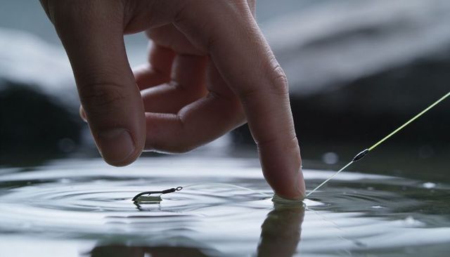 Hand touching water surface with fishing line and hook creating ripples calm reflection