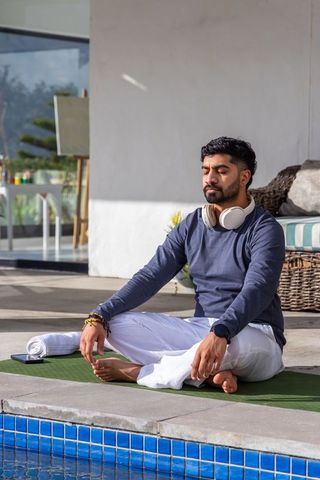 Sunlit asian man meditating poolside with headphones and smartphone for wellness branding