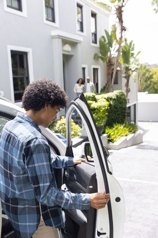 African american man opening car door in suburban driveway while family watches on porch