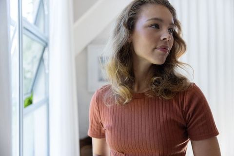 Young woman gazing out window in sunlit minimal interior wearing rust ribbed top