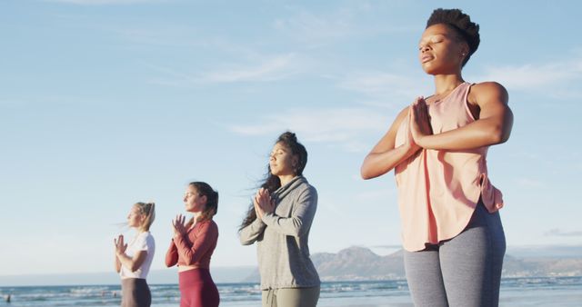 Diverse group of women practicing yoga and meditation on a beach with mountains in the background. Useful for promoting wellness, fitness, meditation programs, women's health, and outdoor activities.