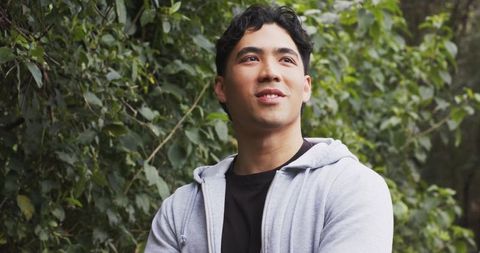 Asian man standing in park wearing light grey hoodie looking upward amid lush greenery