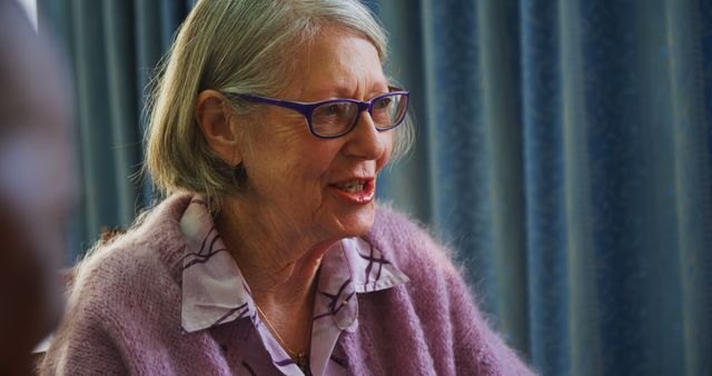 A senior Caucasian woman is smiling warmly, wearing glasses and a cozy pink cardigan, with copy space. Her joyful expression suggests a moment of happiness or a pleasant conversation.