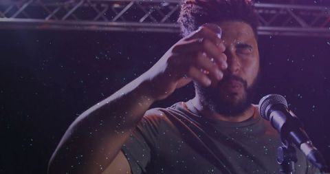 Bearded vocalist wiping face under purple stage lights with microphone and stage particles
