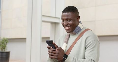 African american man smiling while checking smartphone outside modern building wearing scarf