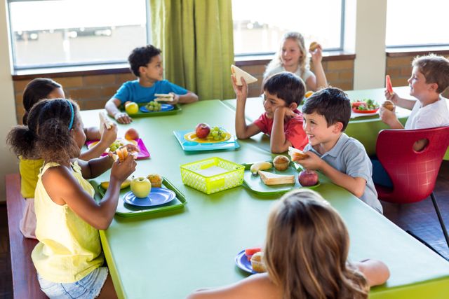Group of diverse schoolchildren enjoying lunch together in a bright cafeteria. They are sitting at a green table, eating healthy meals, and engaging in lively conversation. Ideal for use in educational materials, school brochures, and articles about childhood nutrition and social development.