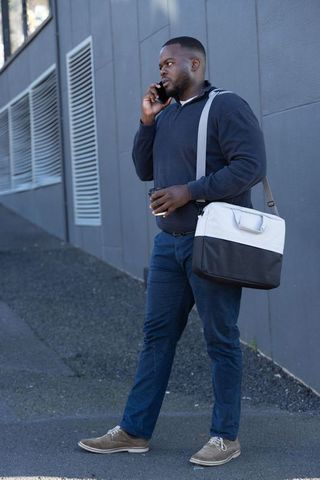 African american commuter walking and talking on phone with coffee and messenger bag