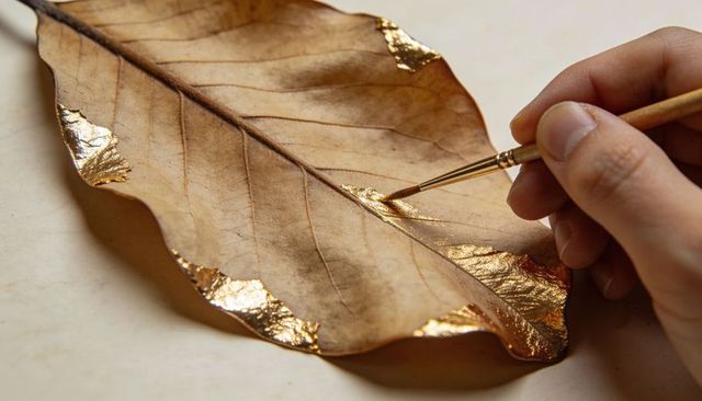 Right hand using brush applying gold leaf to dried leaf for gilded craft detail